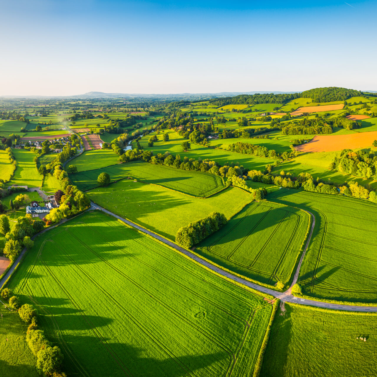 Aerial panorama over picturesque river valley meandering between rolling hills of patchwork pasture, agricultural crops, rural homes and green summer landscape.